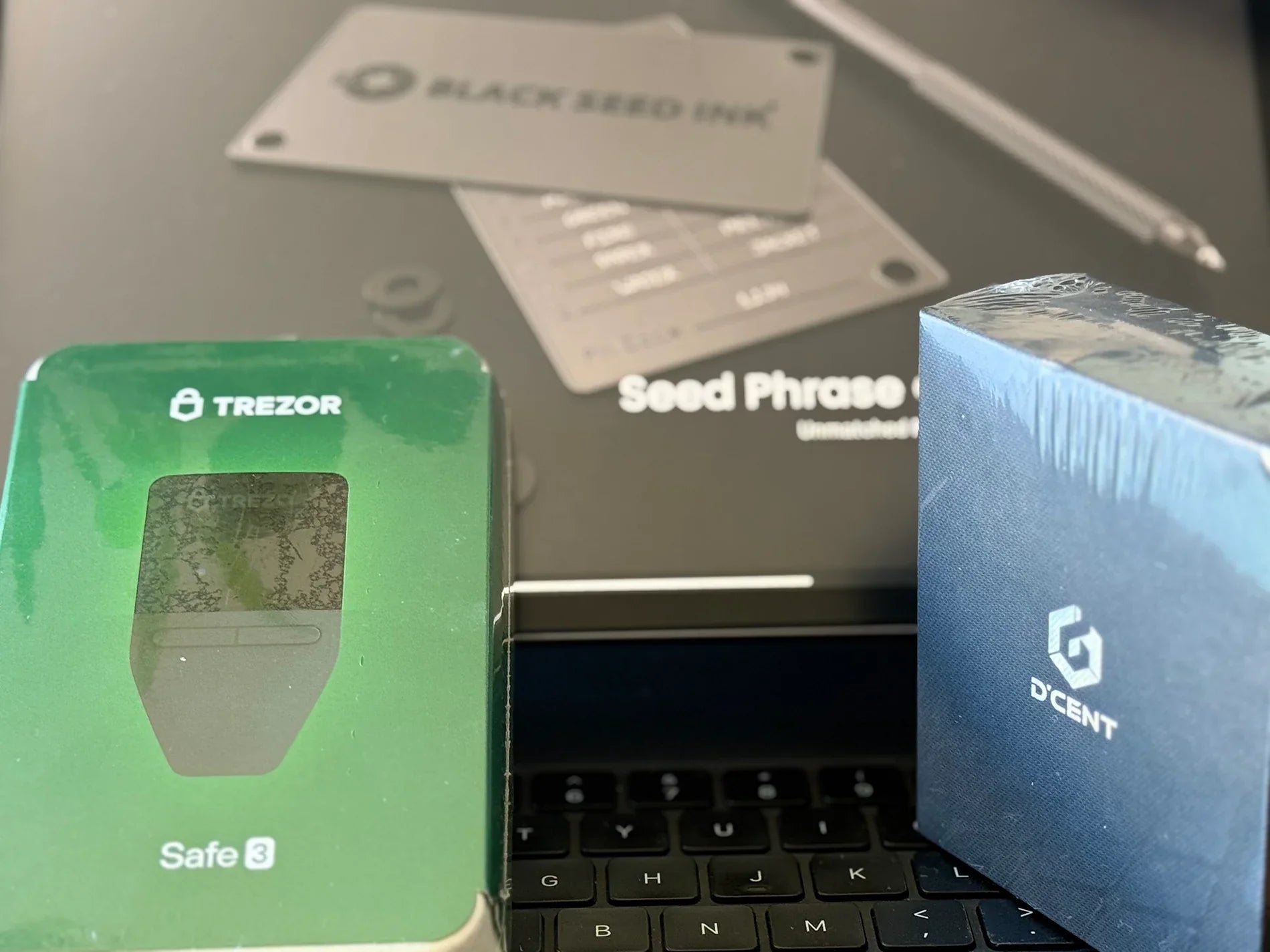 Trezor and D'Cent wallets in packaging atop a iPad keyboard and iPad
