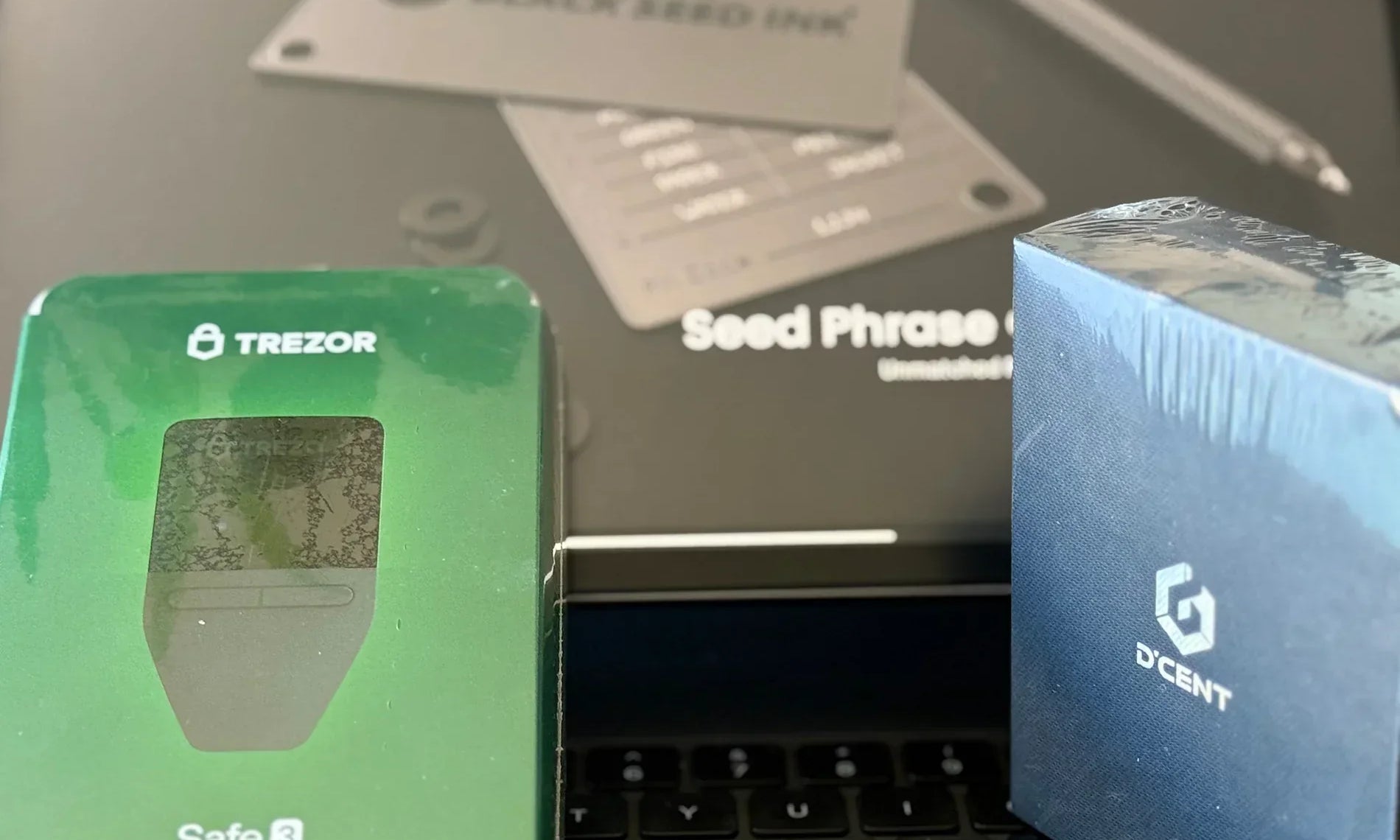 Trezor and D'Cent wallets in packaging atop a iPad keyboard and iPad